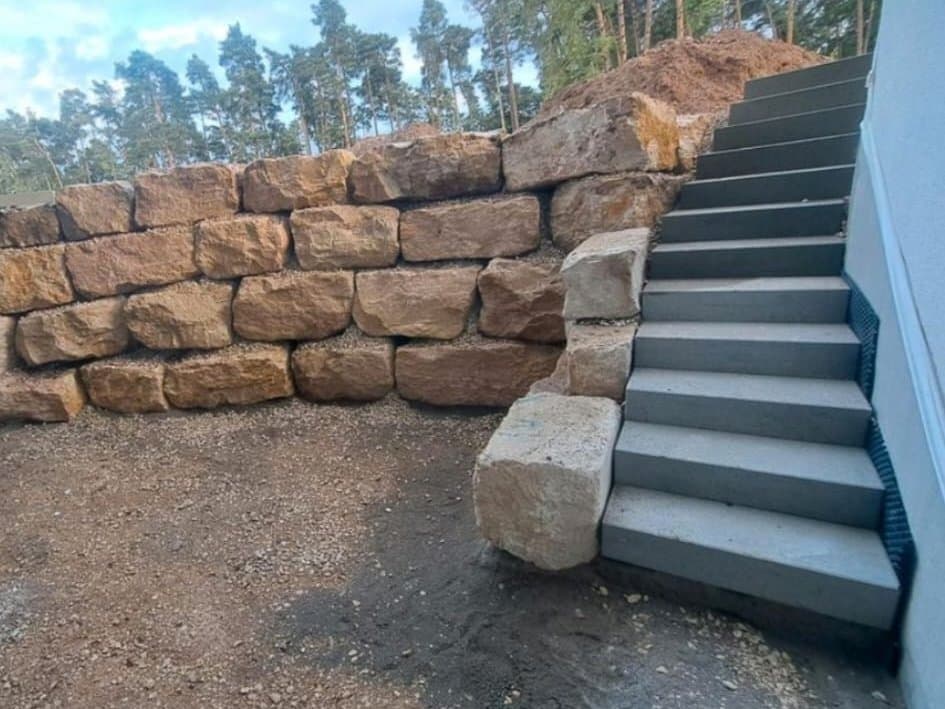 Trockenmauer aus geschichteten Sandsteinquadern, die an eine Außentreppe mit grauen Stufen anschließt